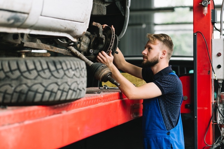 A mechanic in blue overalls is repairing the undercarriage of a vehicle on a red hydraulic lift in an automotive body shop.