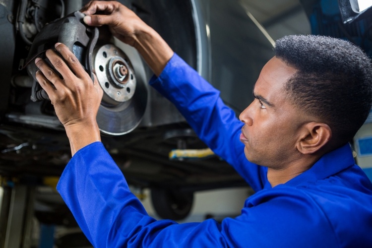 A mechanic wearing a blue jumpsuit is meticulously examining and working on a car's brake system, possibly even conducting a brake pad replacement, in an auto repair shop.